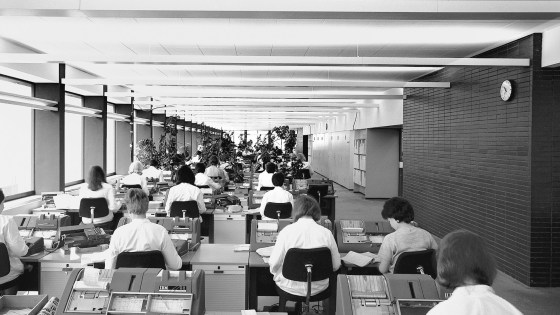 pohled do dlouhé kancelářské místnosti s pracovníky sedícími u počítačů