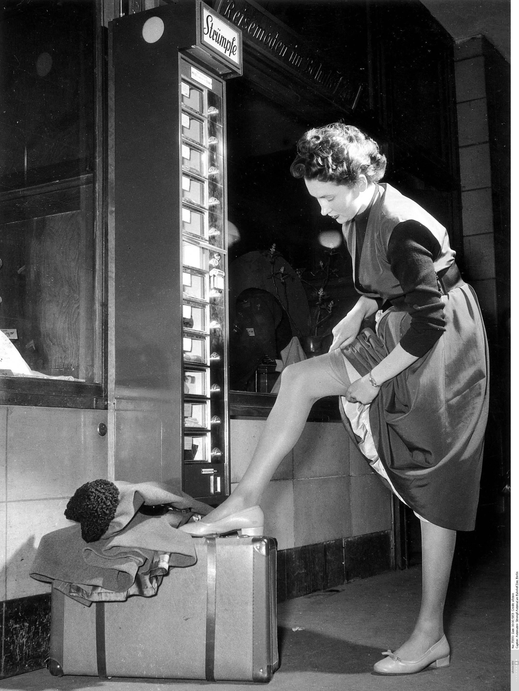 žena si na svou levou nohu podloženou cestovním kufrem navléká punčochu, pravděpodobně zakoupenou v blízkém automatu na punčochové zboží