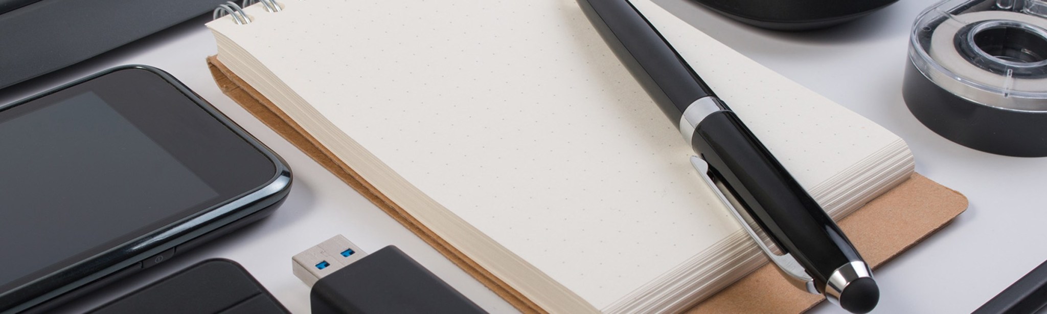 pohled na desku pracovního stolu v kanceláři: telefon, nabíječka, notes s propiskou, lepicí páska